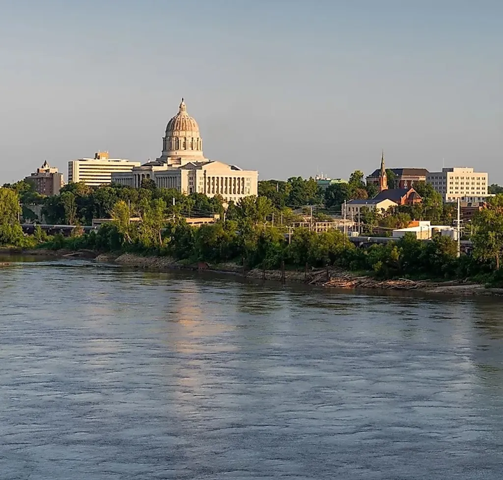 The Missouri State Capitol building with its golden dome rises above Jefferson City, viewed across the Missouri River at golden hour.